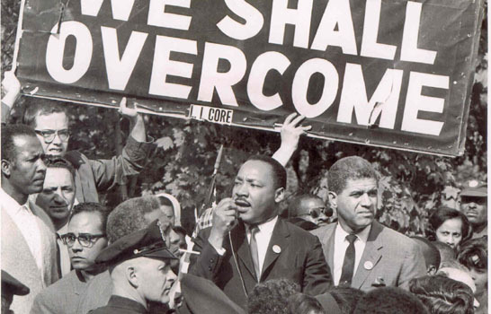 Martin Luther King speaking at a civil rights rally under a sign reading 'We Shall Overcome'